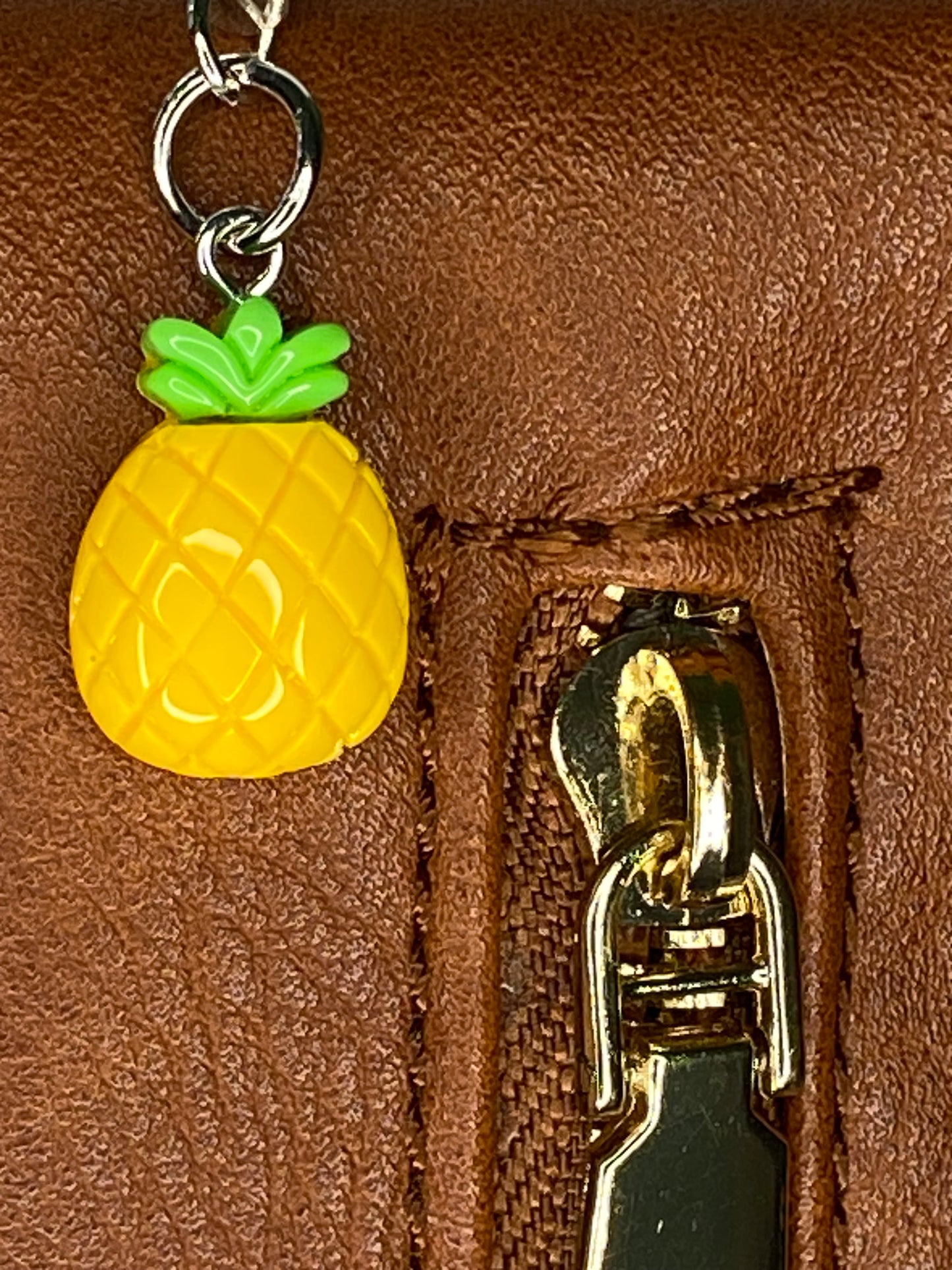 Close-up of a pineapple-shaped keychain attached to a brown leather bag with a gold zipper.